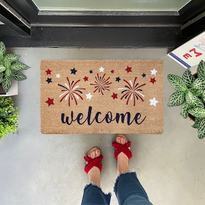 May include: A welcome mat with a red, white, and blue firework design and the word "welcome" in navy blue. The mat is made of coir and has a natural brown color.