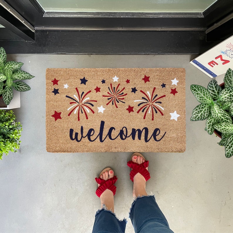 May include: A welcome mat with a red, white, and blue firework design and the word "welcome" in navy blue. The mat is made of coir and has a natural brown color.