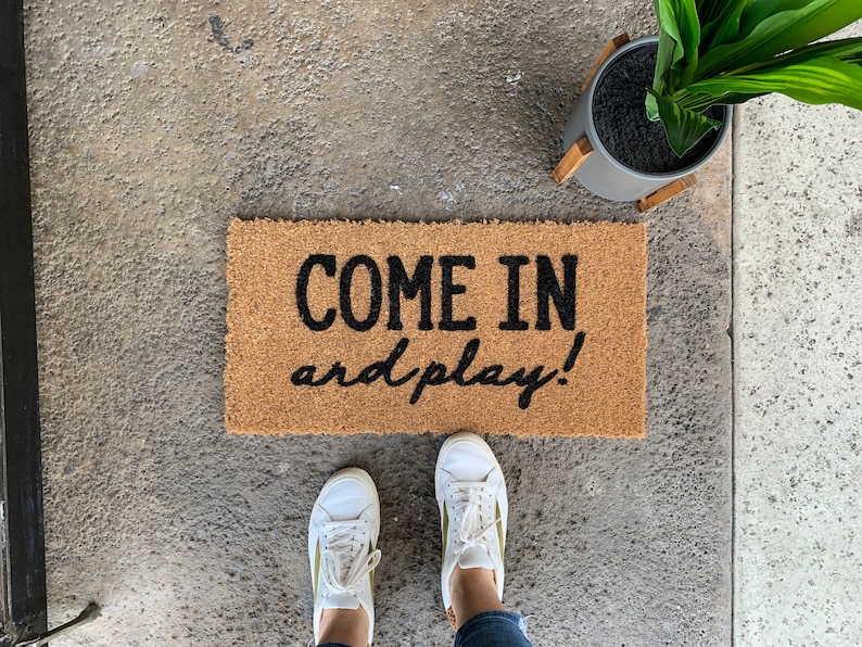 May include: A brown coir doormat with the text "COME IN and play!" in black lettering.