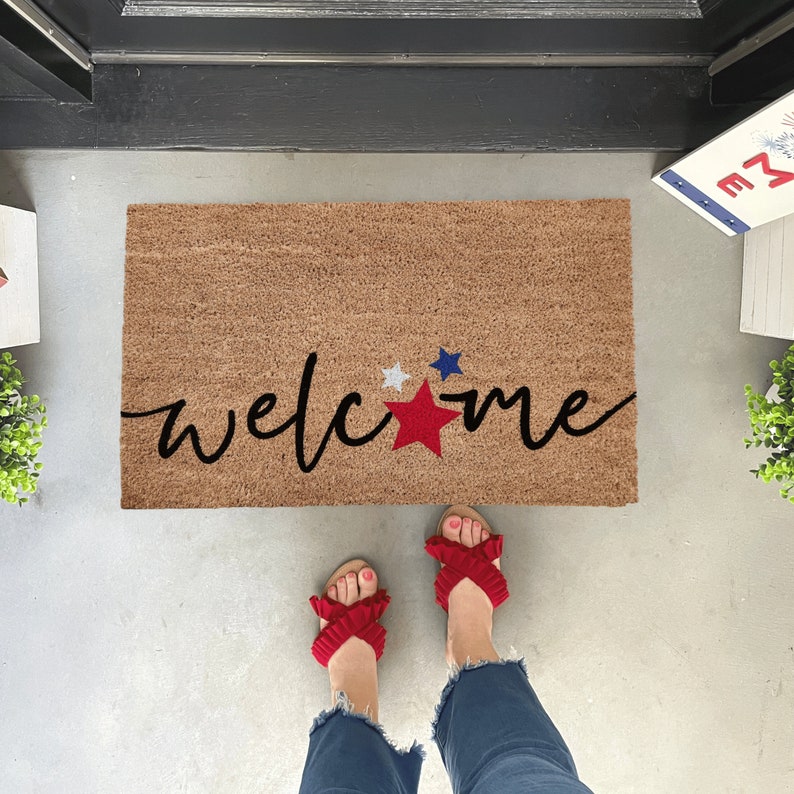 May include: A welcome mat with the word "welcome" written in black script with red, white, and blue stars. The mat is brown and has a rough texture.