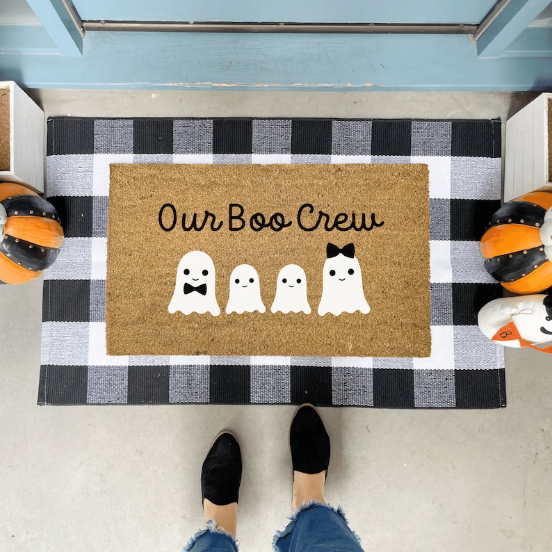 May include: A brown coir doormat with the text "Our Boo Crew" in black lettering. Four white ghost figures with black facial features are arranged in a row below the text. The doormat is on a black and white checkered rug.