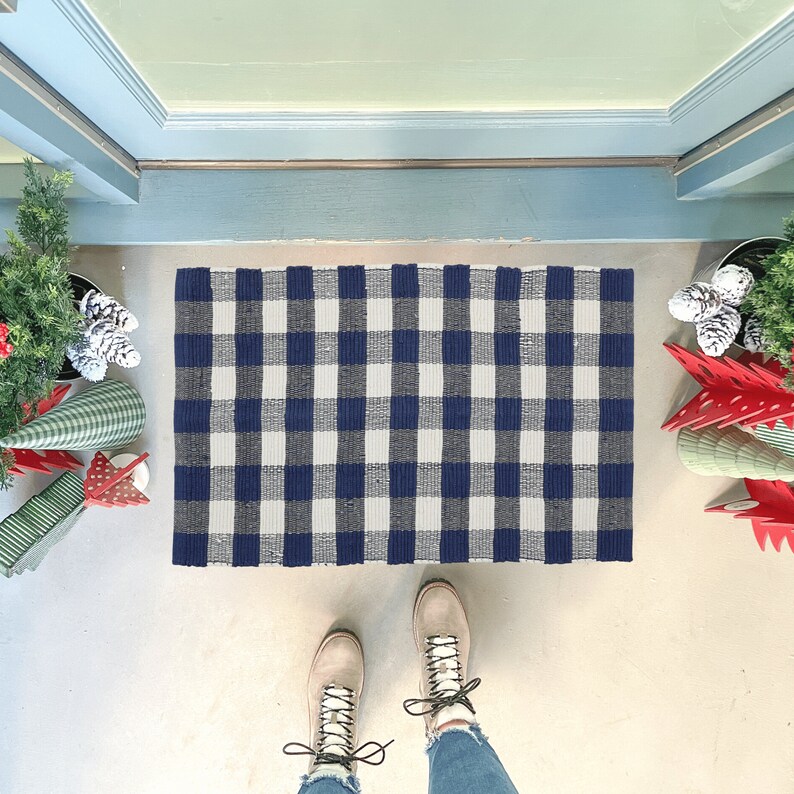 May include: A rectangular doormat with a navy blue and white checkered pattern. The mat is placed in front of a blue door. The image also shows a person's feet wearing light brown boots and decorative Christmas trees.