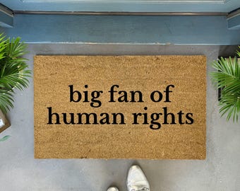 May include: A rectangular doormat made of natural fibers with the phrase "big fan of human rights" in black lettering. The mat is placed on a concrete surface, with a pair of silver shoes visible at the bottom.