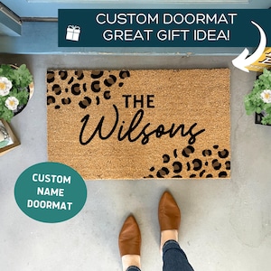 May include: A rectangular coir doormat with a leopard print design in the corners and the family name "The Wilsons" in black script. The text "CUSTOM DOORMAT GREAT GIFT IDEA!" is above the mat. The text "CUSTOM NAME DOORMAT" is on a teal circle.