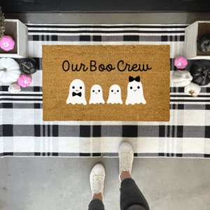 May include: A brown coir doormat with the text "Our Boo Crew" in black lettering. Four white ghost figures with black facial features are arranged in a row. The ghost in the middle has a black bow on its head.