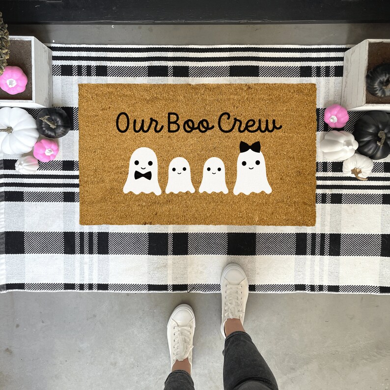 May include: A brown coir doormat with the text "Our Boo Crew" in black lettering. Four white ghost figures with black facial features are arranged in a row. The ghost in the middle has a black bow on its head.
