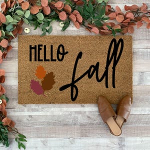 May include: A brown coir doormat with the text "HELLO fall" in black lettering. The text "fall" is in a cursive font. There are three stylized orange, brown, and pink leaves in the upper left corner of the doormat.