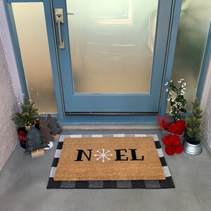May include: A brown coir doormat with a black and white checkered border. The doormat has the word "NOEL" written in black with a white snowflake in between the "N" and "E".