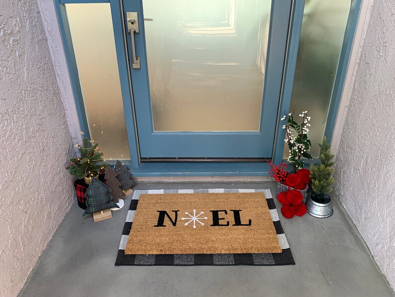 May include: A brown coir doormat with a black and white checkered border. The doormat has the word "NOEL" written in black with a white snowflake in between the "N" and "E".