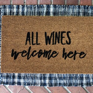 May include: A rectangular doormat with the text "ALL WINES welcome here" in black script. The mat has a brown coir center and a blue and white plaid border, perfect for a home entrance.