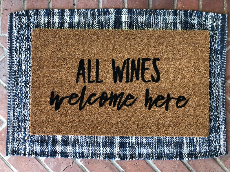 May include: A rectangular doormat with the text "ALL WINES welcome here" in black script. The mat has a brown coir center and a blue and white plaid border, perfect for a home entrance.