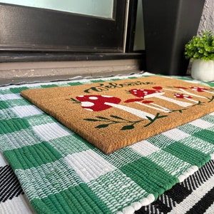 May include: A brown coir doormat with the word "Welcome" and a design of red and white mushrooms. The doormat is on top of a green and white checkered rug.