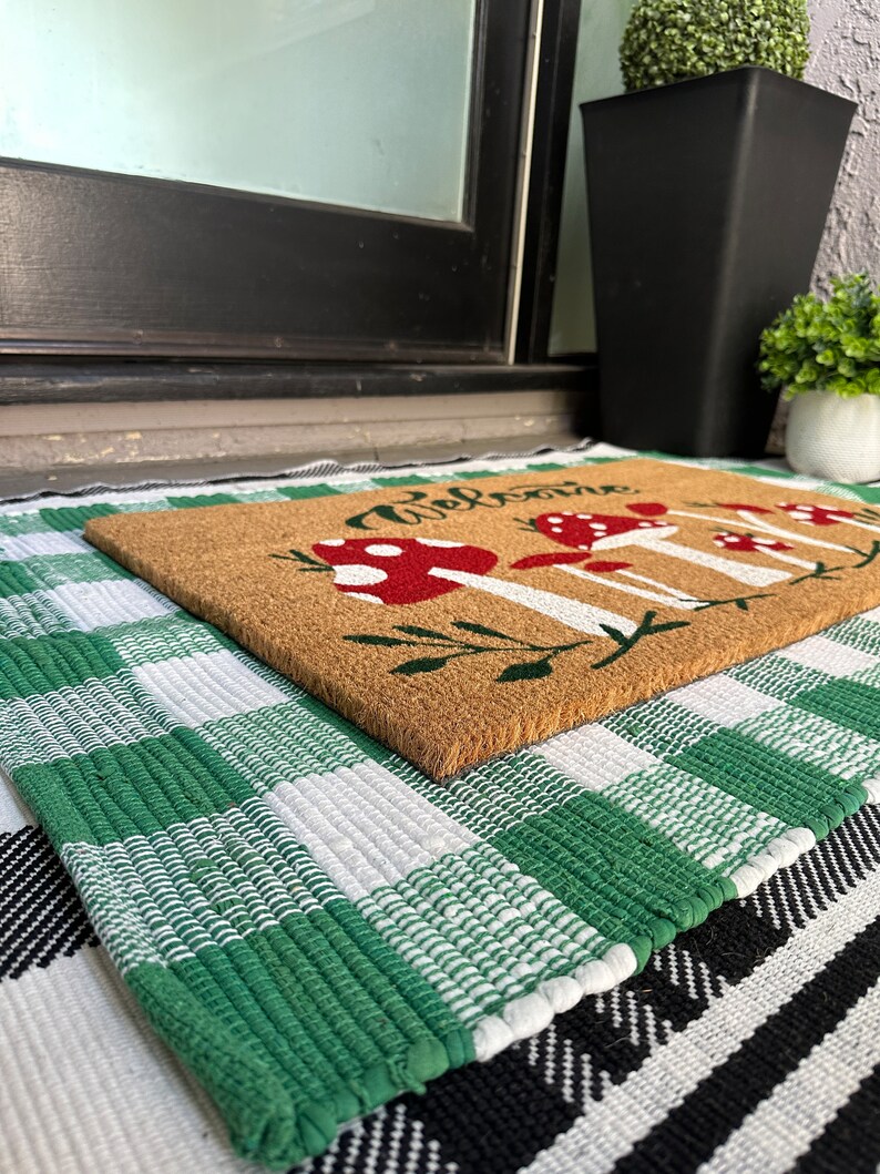 May include: A brown coir doormat with the word "Welcome" and a design of red and white mushrooms. The doormat is on top of a green and white checkered rug.