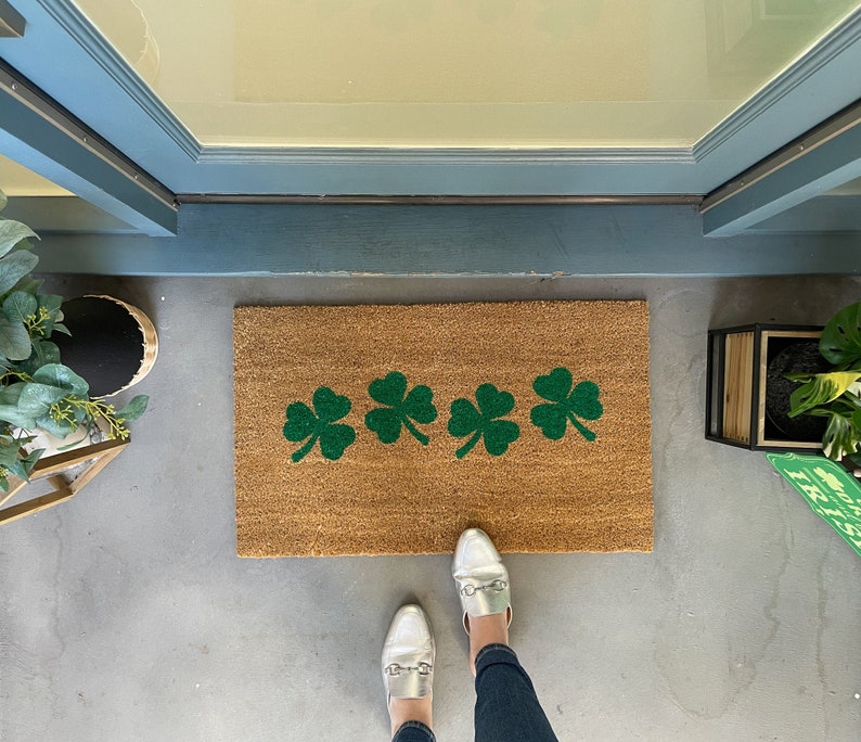 May include: A brown doormat with green shamrock designs. The doormat is on a concrete floor with a pair of white shoes on top of it.
