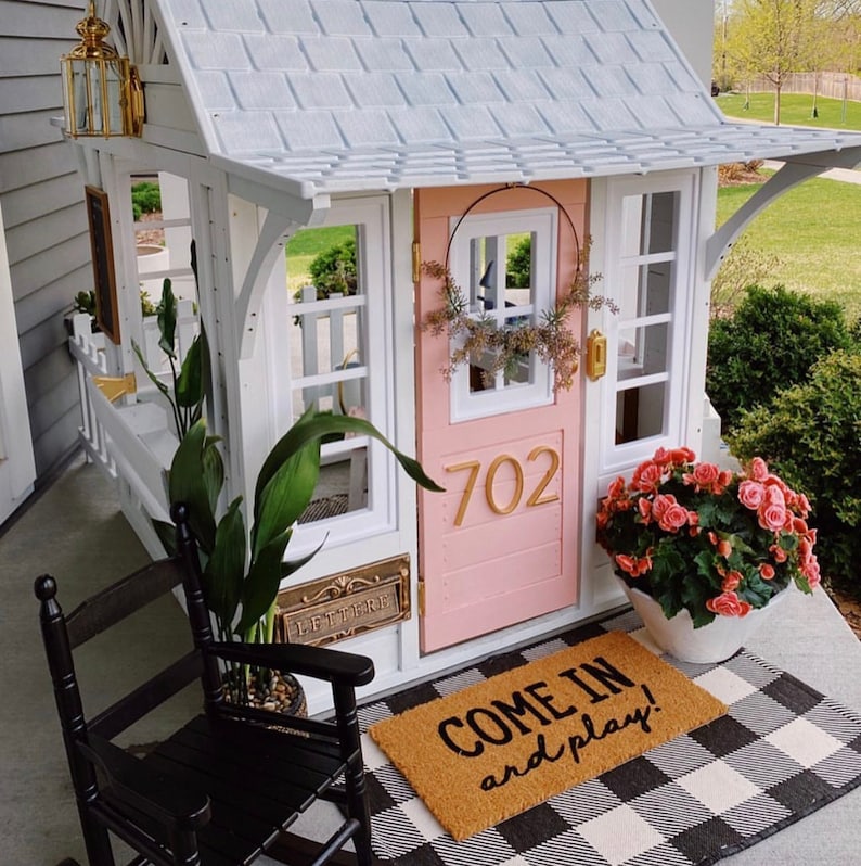 May include: A white and pink playhouse with a black and white chequered rug, a rocking chair, and a welcome mat that says "Come in and play!"