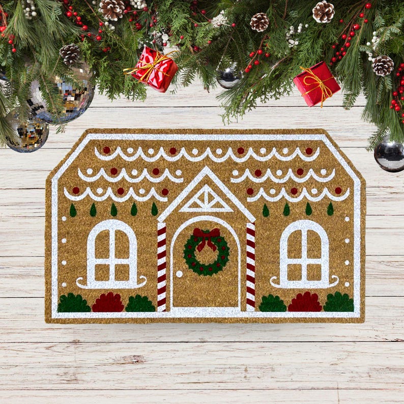 May include: A brown coir doormat shaped like a gingerbread house with white icing and red candy details. The doormat has a wreath on the door and two windows.