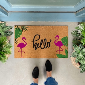 May include: A brown coir doormat with the word "hello" in black script. Two pink flamingos with green palm leaves are on either side of the word.