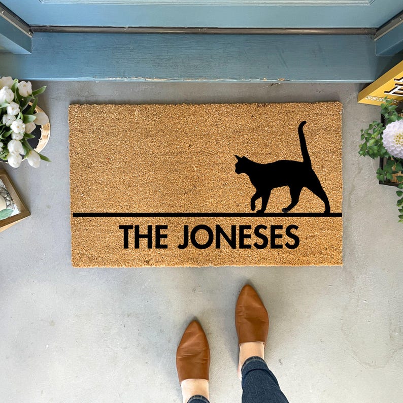 May include: A brown coir doormat with a black silhouette of a cat walking across the top. The text "THE JONESES" is printed in black below the cat.