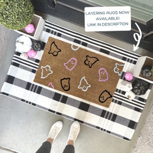 Pode incluir: Um tapete xadrez preto e branco com um tapete de coco marrom com um design de fantasma. O tapete tem contornos de fantasmas rosa e azul. O texto "LAYERING RUGS NOW AVAILABLE! LINK IN DESCRIPTION" &eacute; vis&iacute;vel na imagem.