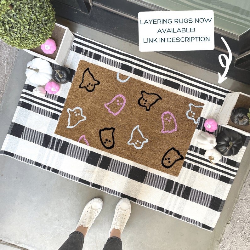 Pode incluir: Um tapete xadrez preto e branco com um tapete de coco marrom com um design de fantasma. O tapete tem contornos de fantasmas rosa e azul. O texto "LAYERING RUGS NOW AVAILABLE! LINK IN DESCRIPTION" &eacute; vis&iacute;vel na imagem.
