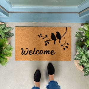 Puede incluir: Una alfombra de coco marr&oacute;n con la palabra "Welcome" en letras cursivas negras. Tres p&aacute;jaros negros est&aacute;n posados en una rama con hojas, tambi&eacute;n en negro, sobre la palabra "Welcome".