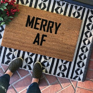 May include: A brown coir doormat with the text "Merry AF" in black lettering. The doormat is on a black and white patterned rug.