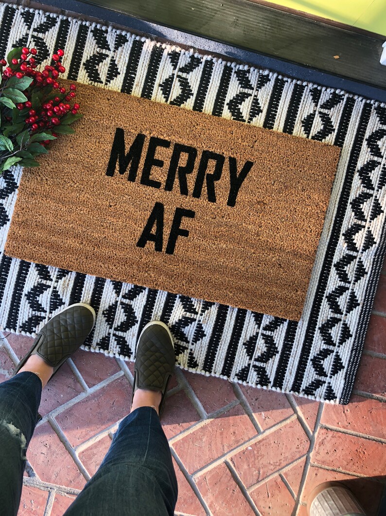 May include: A brown coir doormat with the text "Merry AF" in black lettering. The doormat is on a black and white patterned rug.