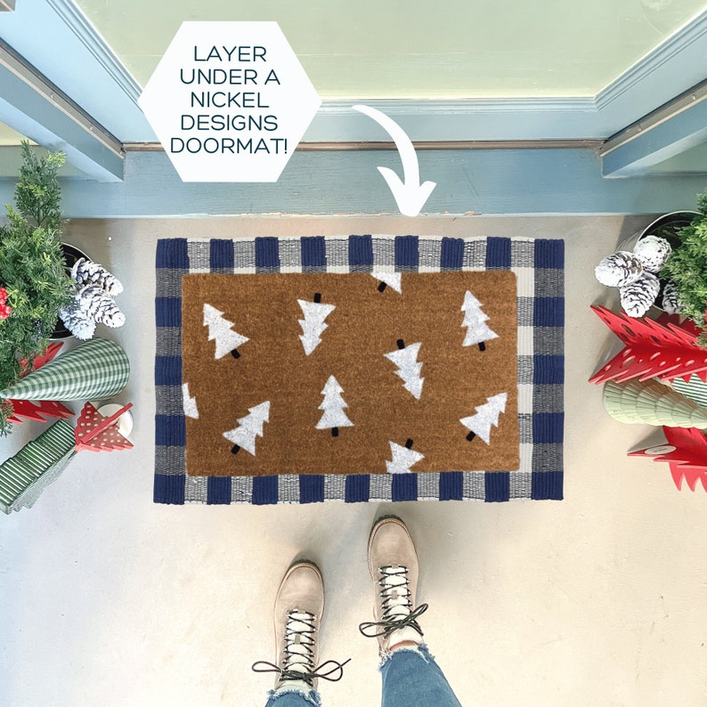 May include: A layered doormat set. The bottom mat is a brown rectangle with white Christmas trees. The top mat has a navy blue and gray checkered border. The text in the image says "LAYER UNDER A NICKEL DESIGNS DOORMAT!"
