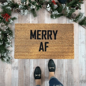 May include: A brown doormat with the text "Merry AF" in black lettering. The doormat is on a light wood floor with a sprig of greenery in the background.