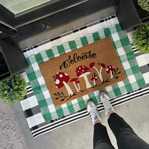 May include: A welcome mat with a green and white checkered pattern. The mat has a brown welcome mat insert with the word "Welcome" and a design of red and white mushrooms.