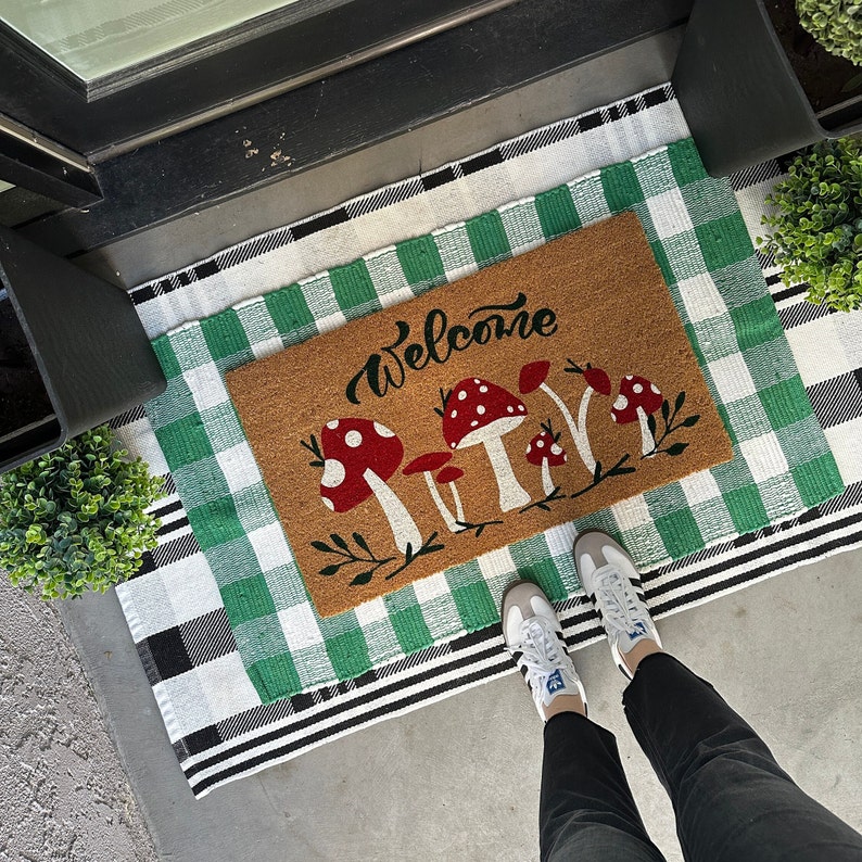 May include: A welcome mat with a green and white checkered pattern. The mat has a brown welcome mat insert with the word "Welcome" and a design of red and white mushrooms.