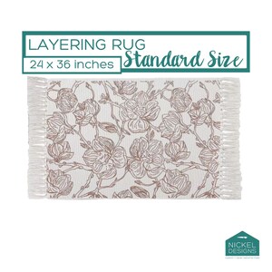 May include: A rectangular layering rug measuring 24 x 36 inches with a white base and a brown floral line art pattern. The rug features fringed edges on the left and right sides. Text on the image reads: LAYERING RUG Standard Size.