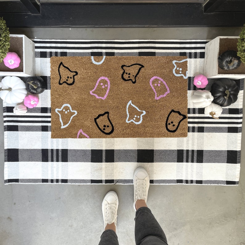 Pode incluir: Um tapete de entrada marrom com um tapete de xadrez preto e branco por baixo. O tapete de entrada tem v&aacute;rias formas de fantasmas pintadas nele em rosa, azul e preto.