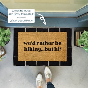 May include: A brown coir doormat with black and white stripes around the edges. The text "we'd rather be hiking...but hi!" is printed in black on the doormat.