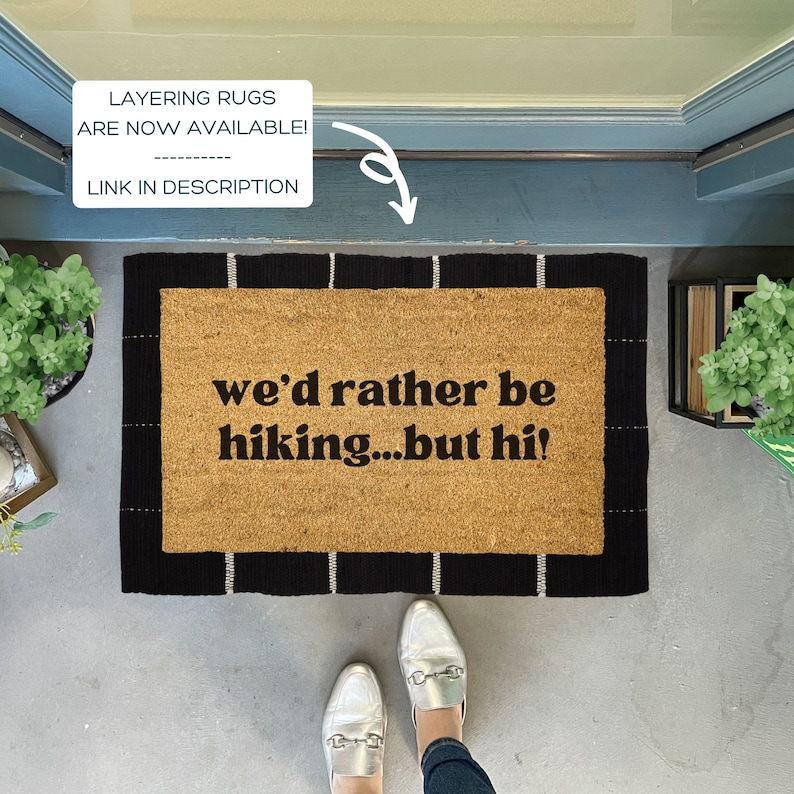 May include: A brown coir doormat with black and white stripes around the edges. The text "we'd rather be hiking...but hi!" is printed in black on the doormat.