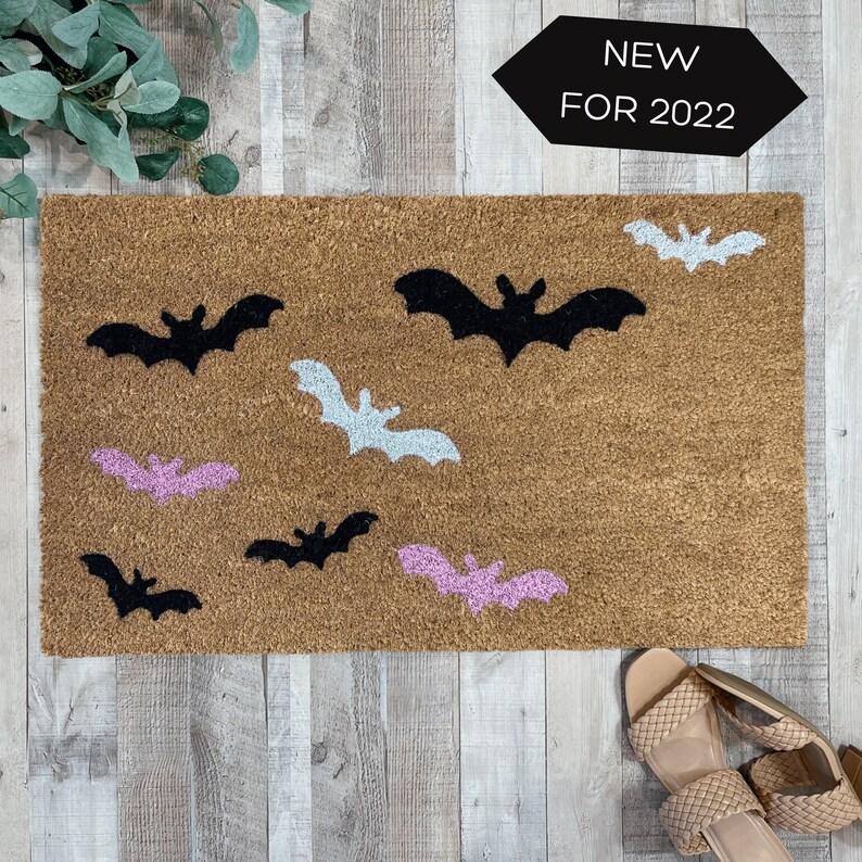 May include: A brown coir doormat with a variety of black and pink bat silhouettes. The text "NEW FOR 2022" is printed on a black hexagonal sign above the doormat.