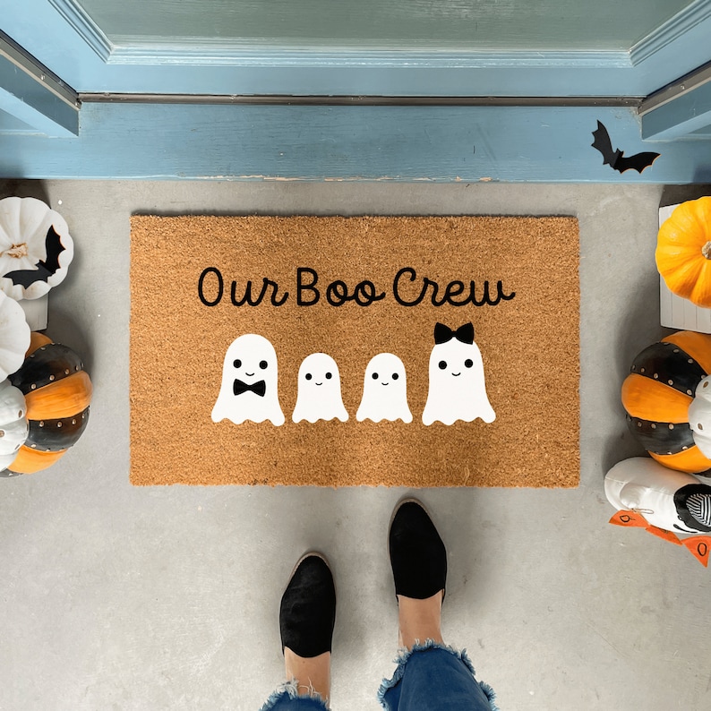 May include: A brown coir doormat with the text "Our Boo Crew" in black script. There are five white ghost figures with black facial features, including a bow on one ghost.