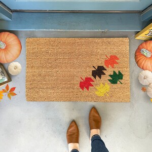Peut inclure: Un paillasson en fibre de coco brun avec un motif de cinq feuilles d'automne colorées dans des tons de rouge, orange, jaune, vert et brun. Le paillasson est sur un sol en béton avec des citrouilles et des courges autour.