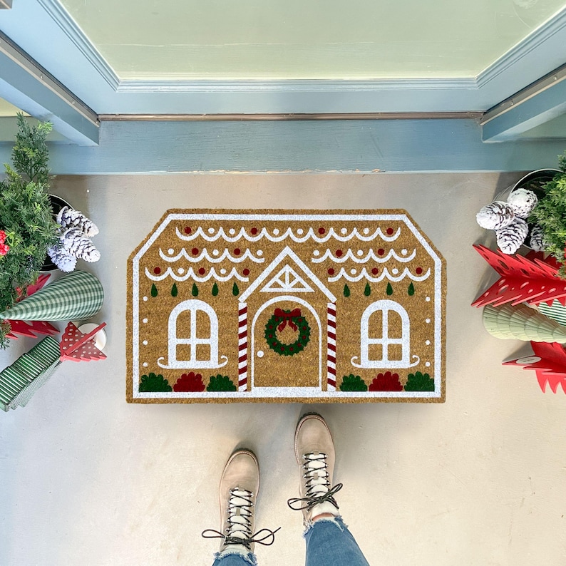 May include: A brown coir doormat shaped like a gingerbread house with white icing and red candy cane accents. The doormat has a wreath on the door and two windows. A pair of brown boots are standing on the doormat.