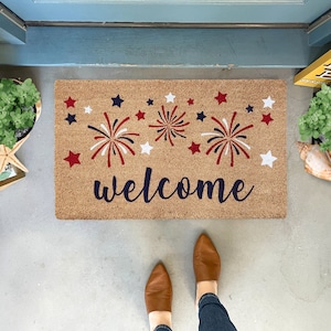 May include: A welcome mat with a brown background and the word "welcome" in navy blue script. The mat is decorated with red, white, and blue fireworks and stars.