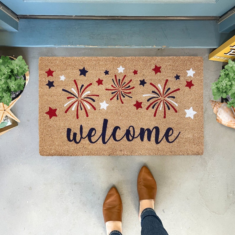 May include: A welcome mat with a brown background and the word "welcome" in navy blue script. The mat is decorated with red, white, and blue fireworks and stars.