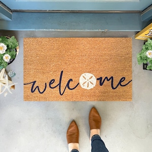 May include: A brown coir welcome mat with the word "welcome" written in navy blue script with a white seashell in the centre.