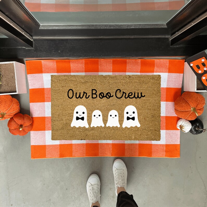 May include: A brown coir doormat with a white and orange checkered border. The doormat has the text "Our Boo Crew" in black lettering and five white ghosts with black bowties.