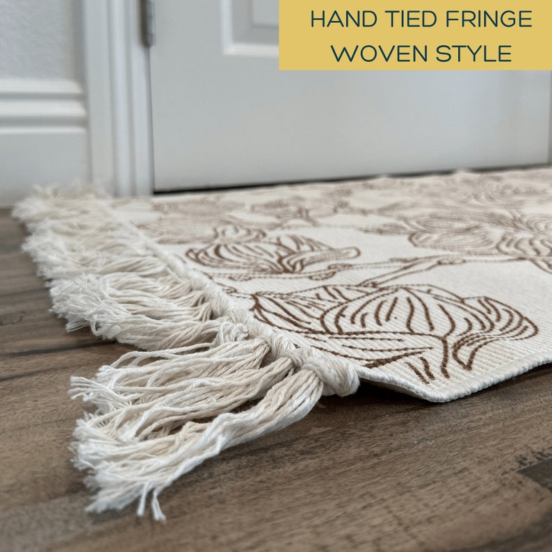 May include: A rectangular woven rug featuring a cream-colored base with a brown floral pattern and hand-tied fringe along the edge. The text HAND TIED FRINGE WOVEN STYLE is visible in a yellow box at the top right of the image.