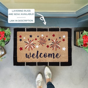 May include: A welcome mat with a brown coir surface and black border. The mat features a red, white, and blue firework design and the word "welcome" in blue.