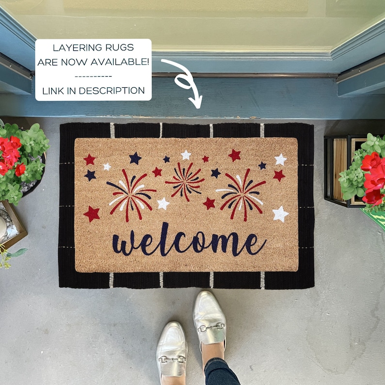 May include: A welcome mat with a brown coir surface and black border. The mat features a red, white, and blue firework design and the word "welcome" in blue.