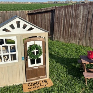 May include: A white and brown wooden playhouse with a green wreath on the door. The playhouse has a sign that says "Come in and play!" and is sitting on a green grassy lawn.