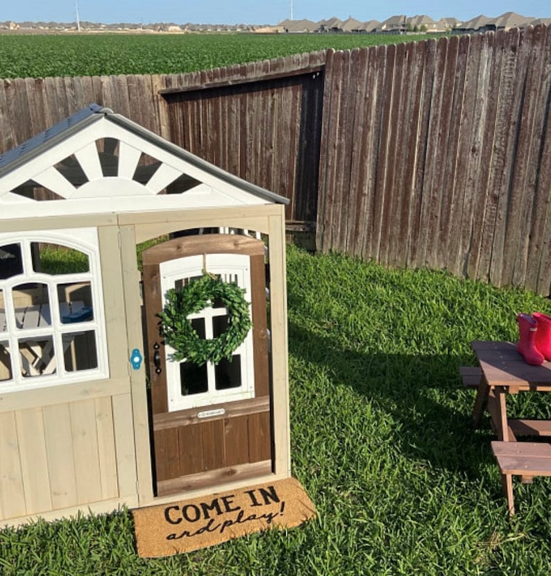 May include: A white and brown wooden playhouse with a green wreath on the door. The playhouse has a sign that says "Come in and play!" and is sitting on a green grassy lawn.