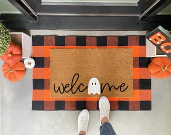 May include: A rectangular tan coir doormat featuring the black text "welcome" with a white ghost replacing the letter "o". The mat is layered over a larger black and orange buffalo plaid rug, creating a decorative entrance display for the home.