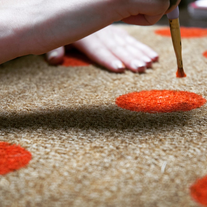 May include: A hand uses a paintbrush to add a red dot to a brown doormat with other red dots.